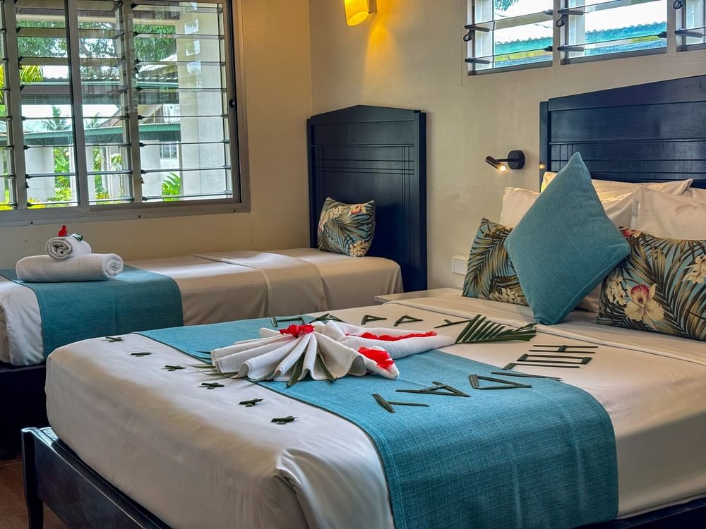 Beds with teal runners and leaf decorations by a window under a warm wall lamp in Family Room at TokaToka Resort Nadi Fiji