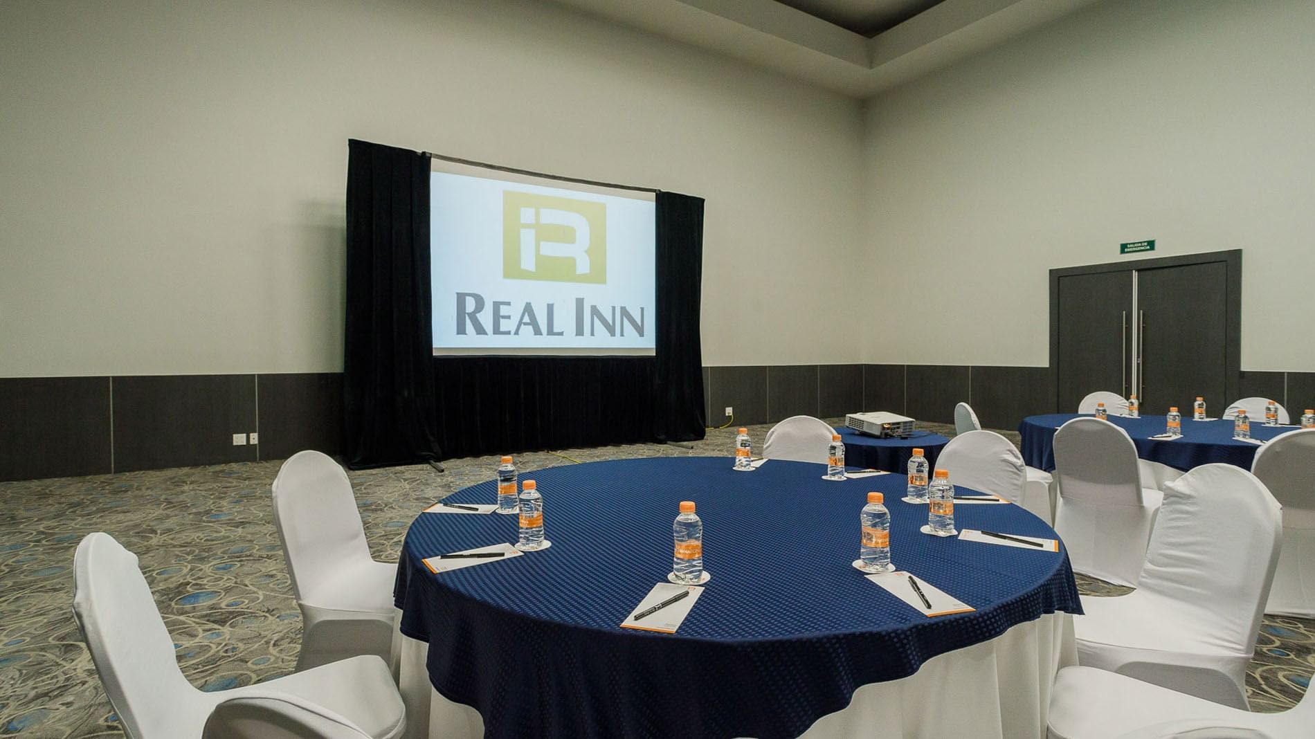 Banquet set-up with blue linens, a projector screen, and a refreshment corner in the Verona Venue at Real Inn Tijuana