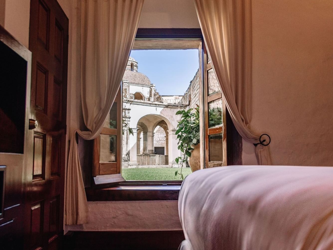 Queen Novicia Suite with beige curtains reveals a historic courtyard through an open window at Quinta Real Oaxaca