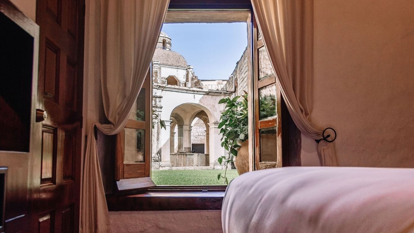 Queen Novicia Suite with beige curtains reveals a historic courtyard through an open window at Quinta Real Oaxaca