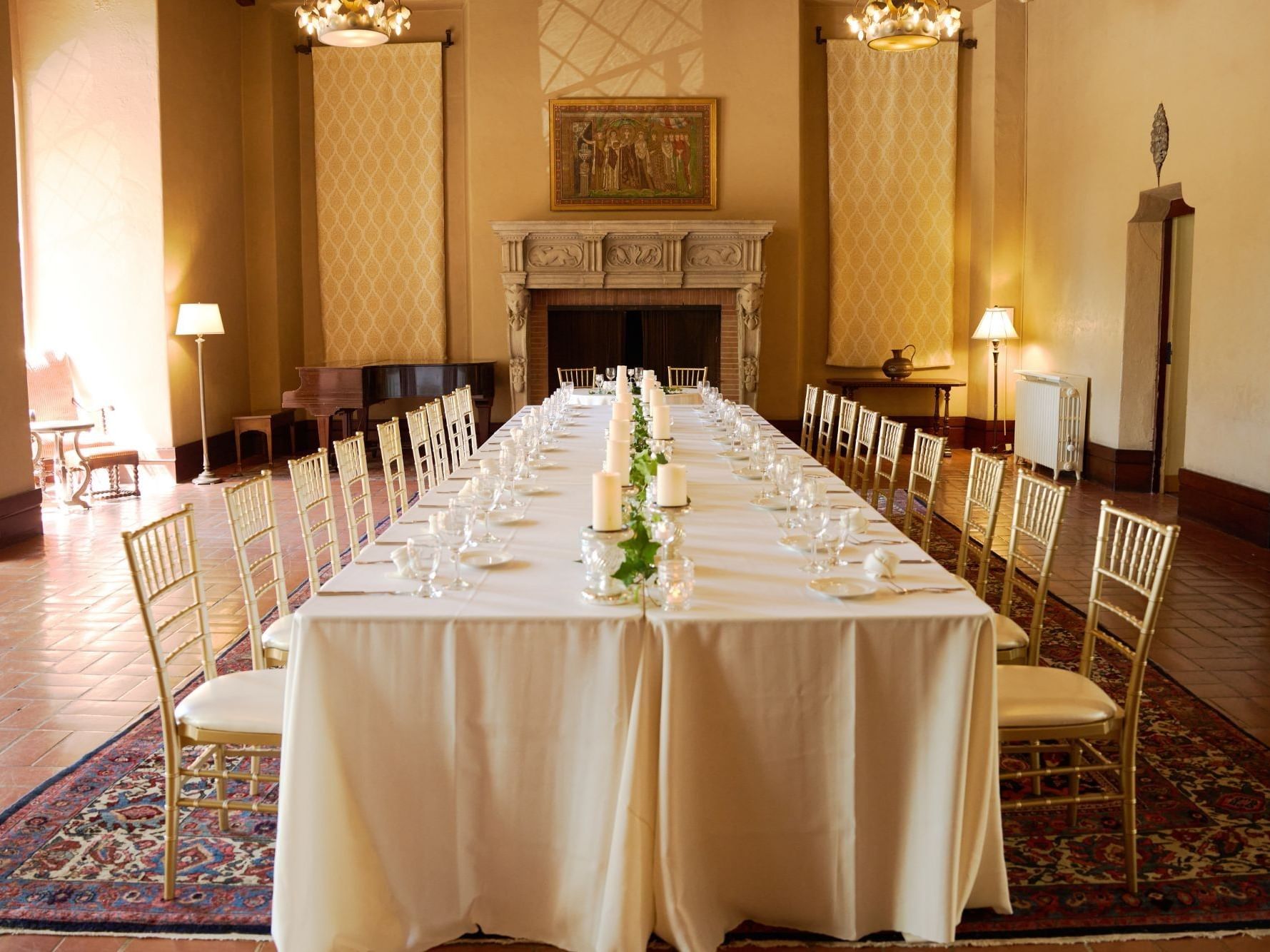 Center table with floor length white linel lined with glassware, ivy, and candle holders. Gold chivalry chairs are on both sides of the table 