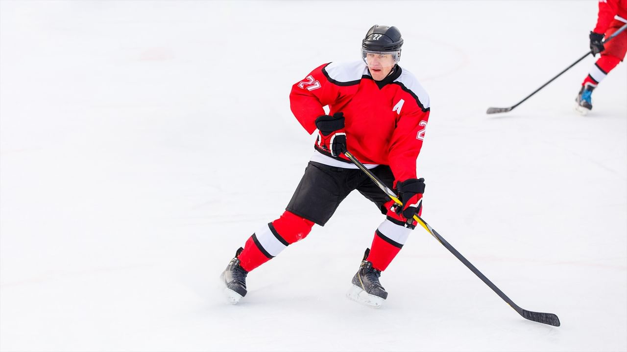 Hockey player in red uniform and black helmet skating on ice holding a hockey stick.