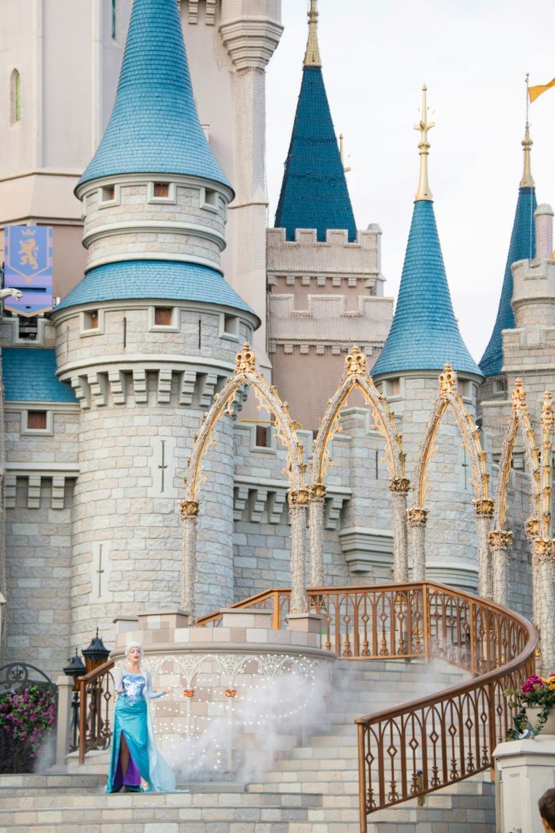 A view of Cinderella Castle at Walt Disney World, with Elsa in her iconic blue dress on the castle steps with a swirling fog effect, part of a performance.