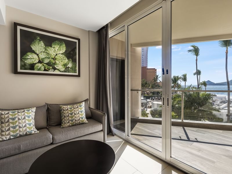 Junior Suite Vista Al Mar living area with a grey sofa, leaf artwork, and a sunny balcony entrance at Sunvivia Mazatlán