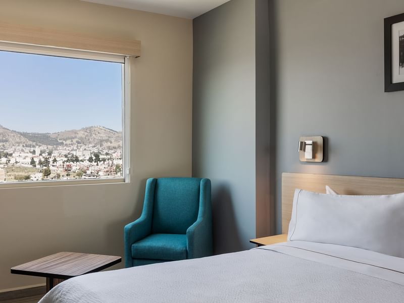 Cozy bed with a blue colored chair and a Window view in Standard King Room at Gamma Hotels