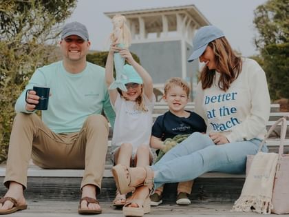 A smiling family of four sits on a wooden deck outdoors with a modern beach house visible in the background.