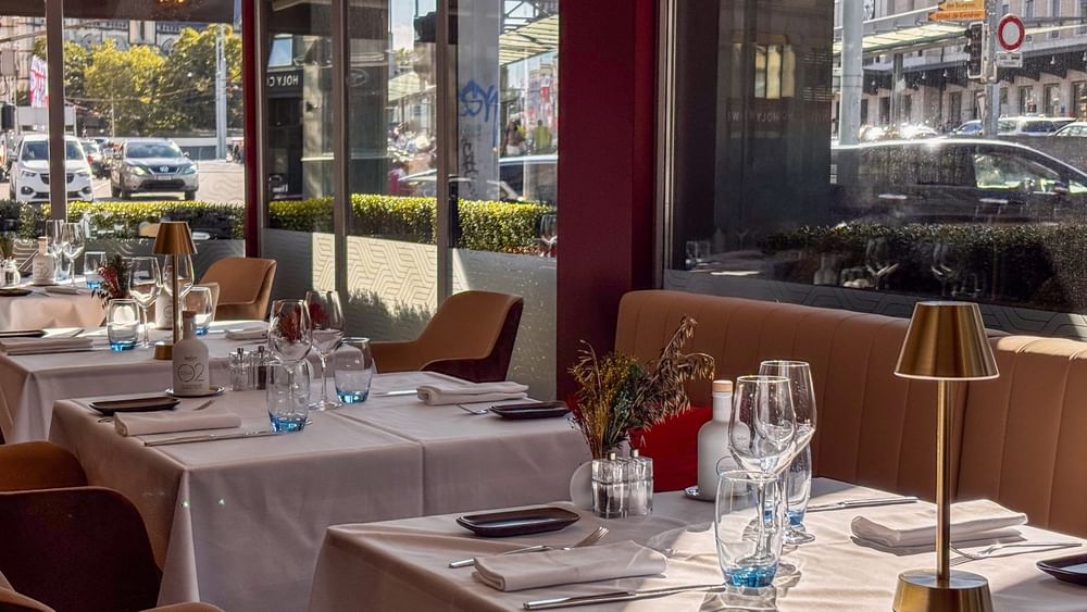 White linen tables with gold lamps by sunlit windows under a red pillar in Téséo at Warwick Geneva