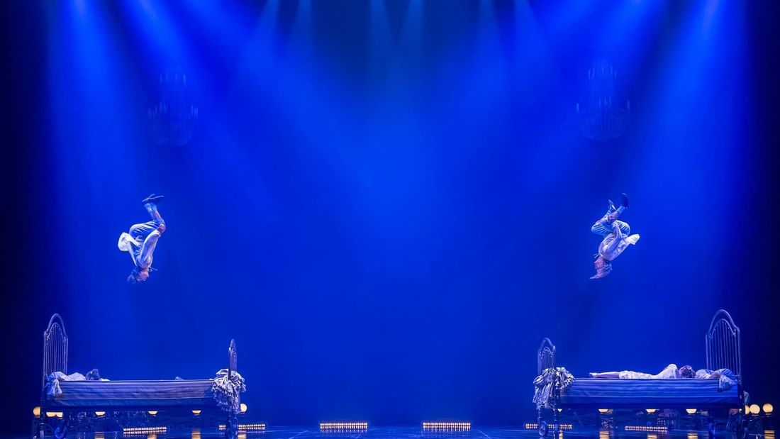 Two performers float above beds on a stage with blue lighting and audience in background.