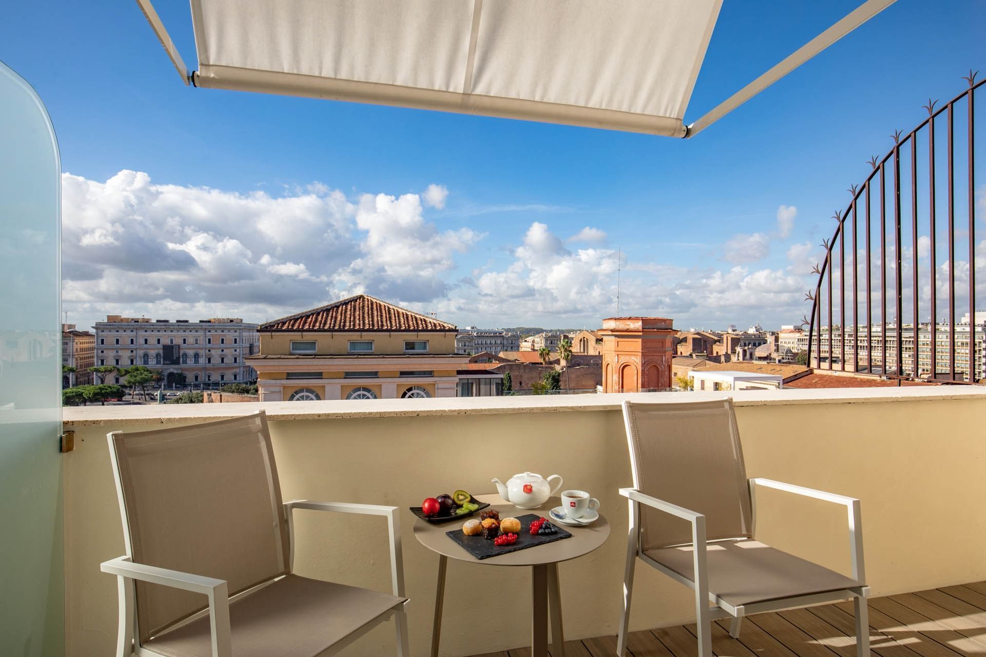 Superior Room with balcony at The Independent, featuring two chairs, a table set with treats, and a cityscape view
