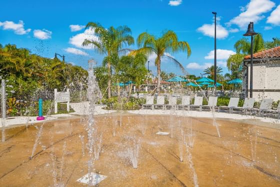 Vista Cay Resort water fountain with multiple jets, surrounded by white lounge chairs and palm trees under a blue sky.