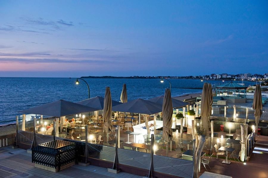 Outdoor seaside terrace with illuminated tables under umbrellas at dusk by the ocean at Le Grand Hôtel des Thermes