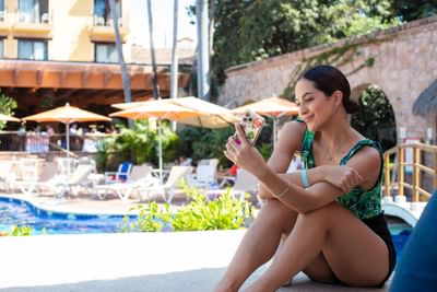 Woman in green top and black shorts, holding a phone, sitting near a pool with umbrellas.