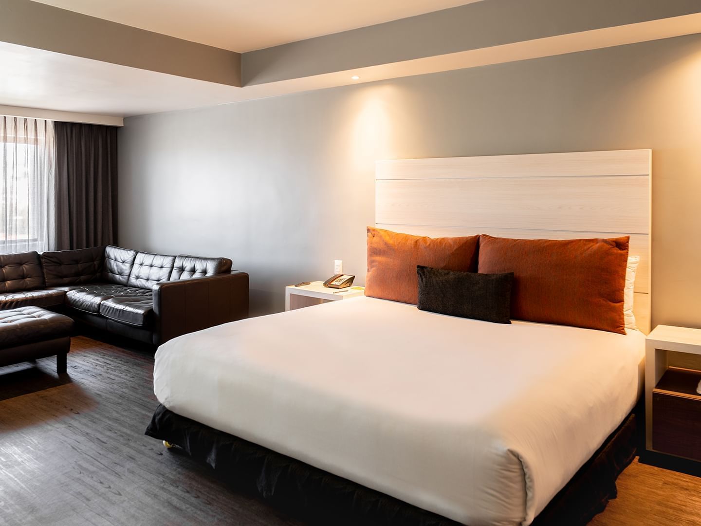 Nightstands & a black leather sectional sofa by the bed in King Junior Suite with dark wood flooring at Real Inn Tijuana