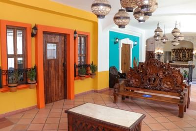 Vestíbulo colorido con paredes amarillas y turquesa, bancas talladas de madera y lámparas colgantes en Los Arcos Suites