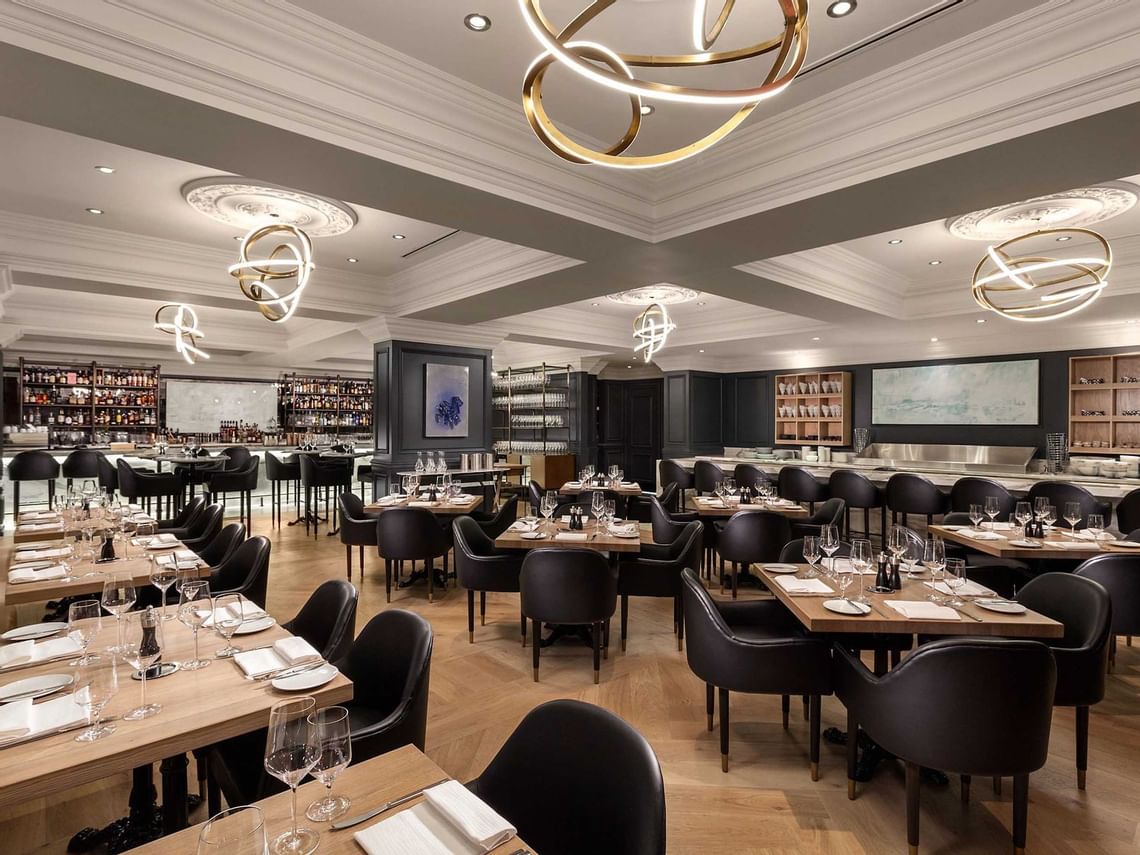 A photo of the Boulevard Kitchen & Oyster Bar dining room interior in Vancouver, Canada. The elegant space features comfortable seating, white tablecloths, and large windows overlooking the city (or another relevant detail if visible). (Photo Credit: The Sutton Place Hotel)