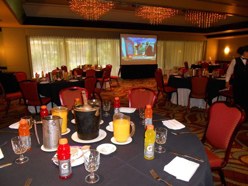 Banquet room with round tables and red chairs facing a large projection screen beneath chandeliers at Miami Lakes Hotel