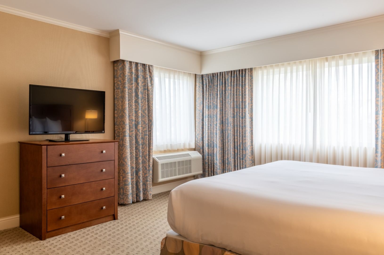 A hotel room with a king-sized bed, a dresser with a TV, and a window with curtains.