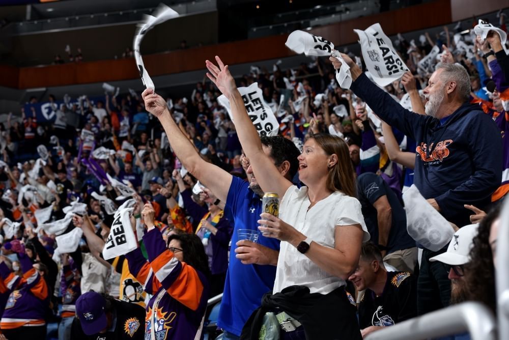 A crowd of cheering people in Solar Bears gear holding drinks and waving white towels that say Let's Go Bears!