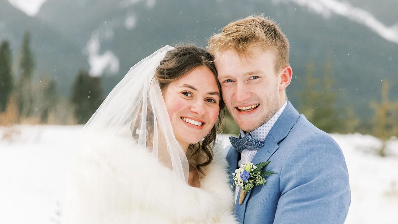A smiling bride and groom in winter attire pose for a photo in a snowy landscape.