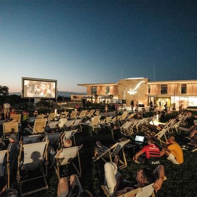 People in chairs watch an outdoor movie on a large screen next to a modern building at dusk.