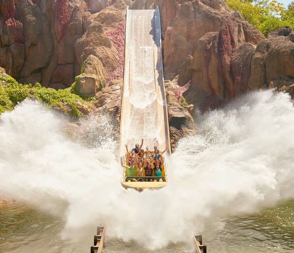 A boat plunges down a steep log flume at a theme park, creating a massive, symmetrical white water splash against rock walls.