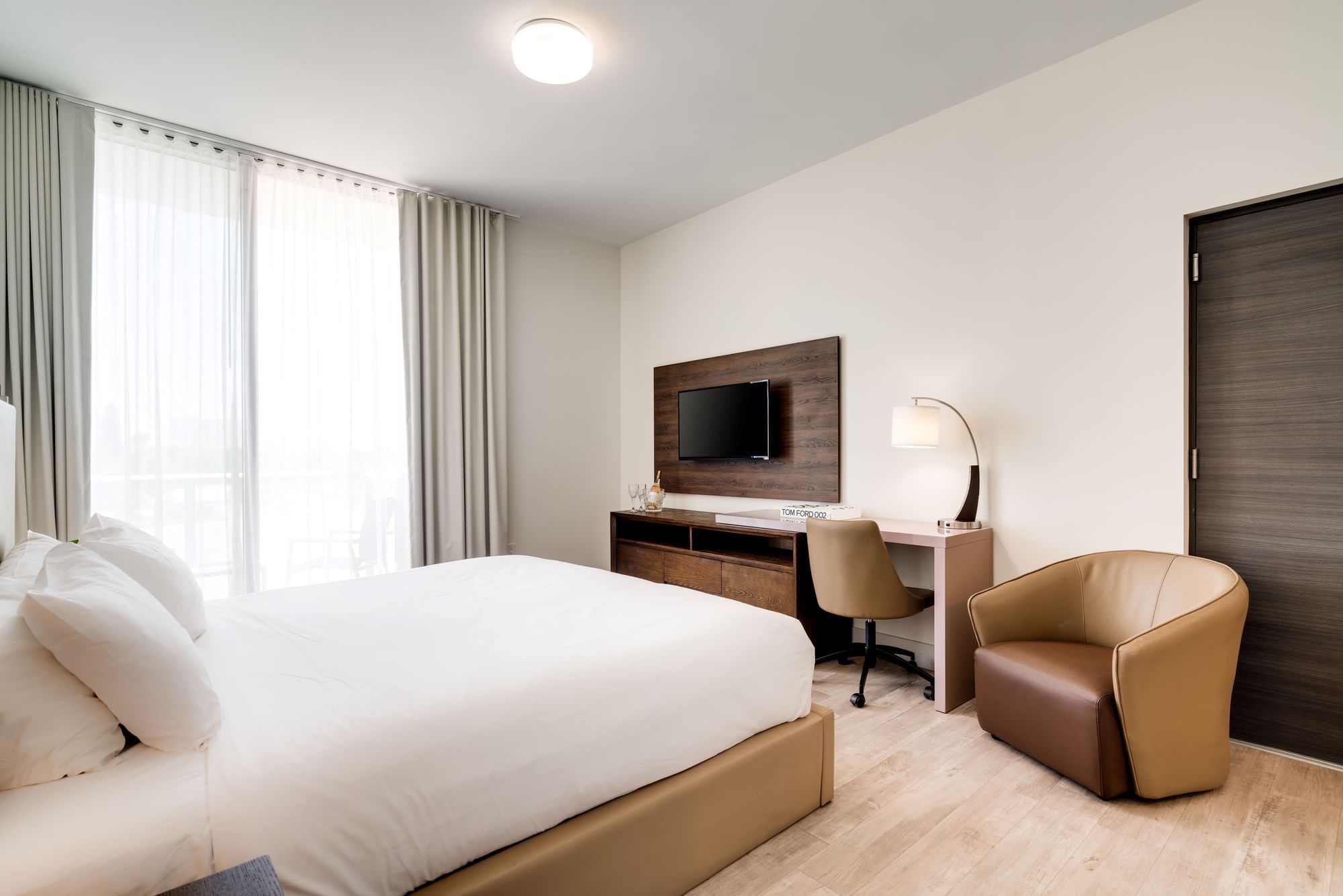A wide-angle view of a hotel room with a large window, comfortable bed, desk with chair, and TV on the wall.