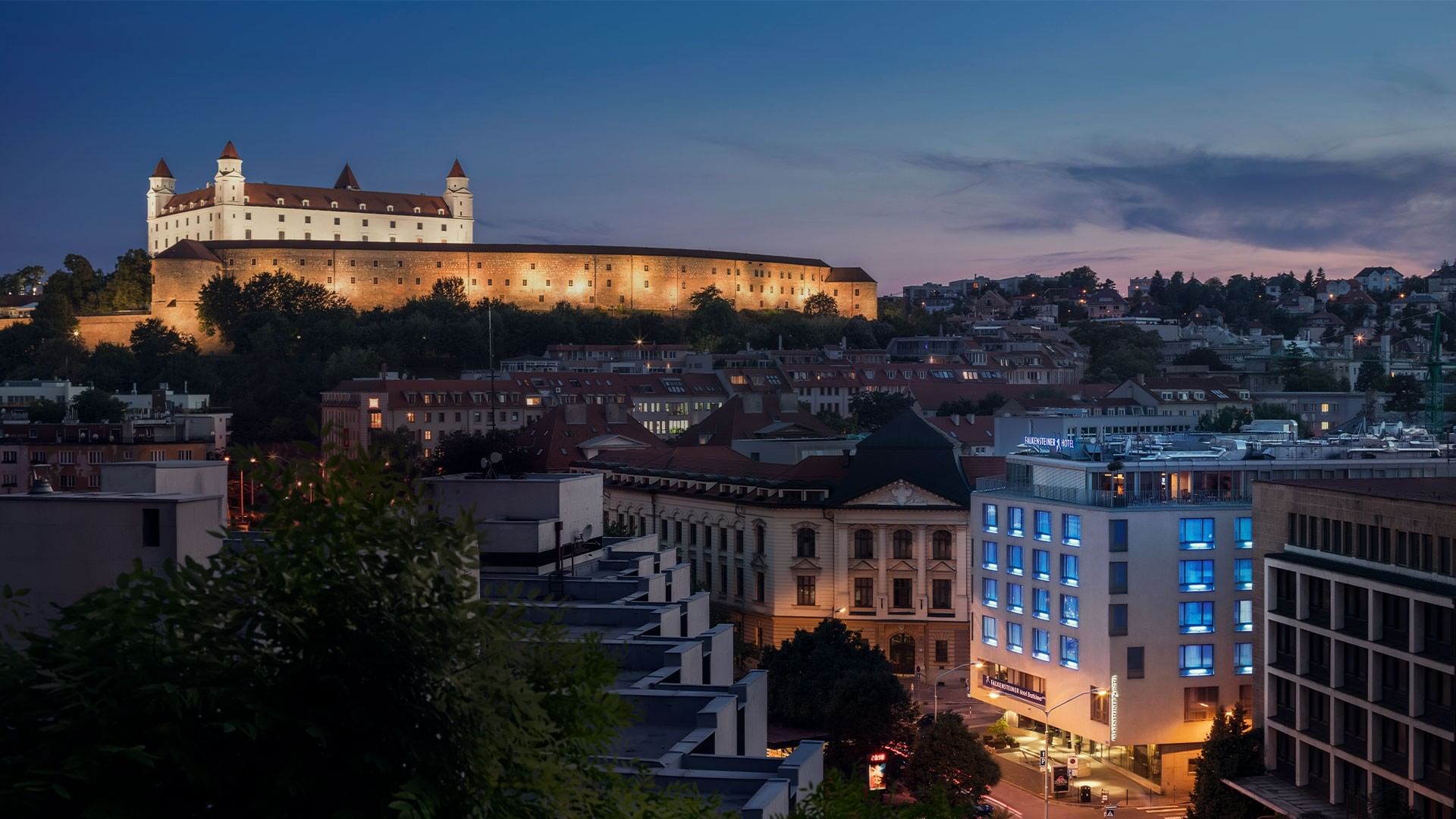 Illuminated Bratislava Castle and cityscape at dusk with modern buildings and a historic castle.