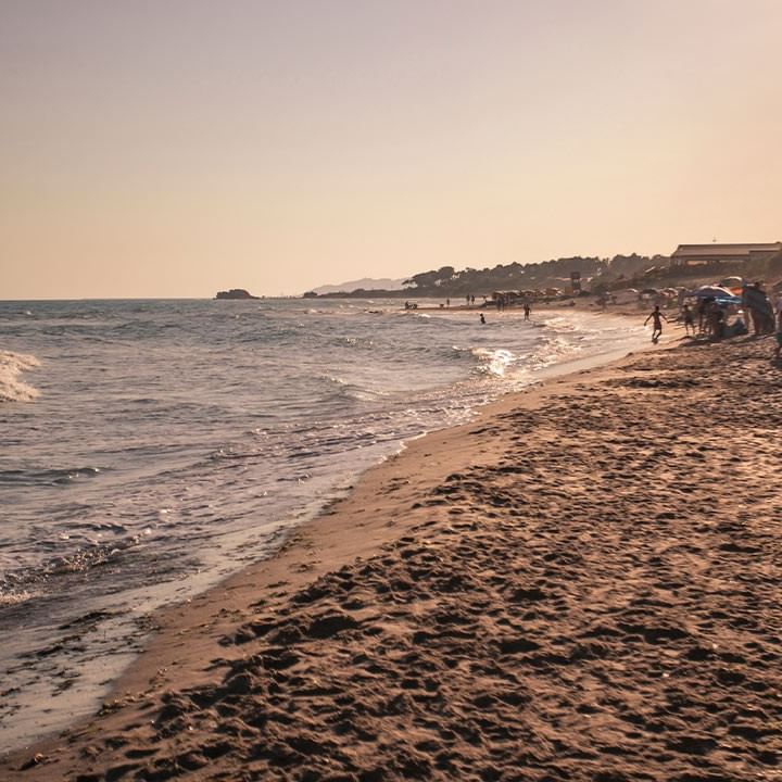 Sandstrand mit Besuchern und Sonnenschirmen, umgeben von Palmen und einem ruhigen Meer bei Sonnenuntergang.