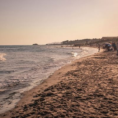 Sandstrand mit Besuchern und Sonnenschirmen, umgeben von Palmen und einem ruhigen Meer bei Sonnenuntergang.