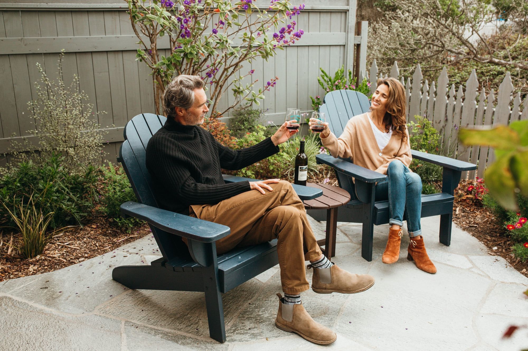 A couple sits in blue Adirondack chairs, toasting with wine glasses in a small outdoor seating area.