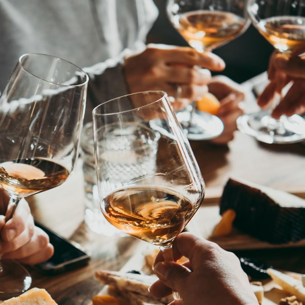 People clink glasses of white wine over a table with cheese and crackers.