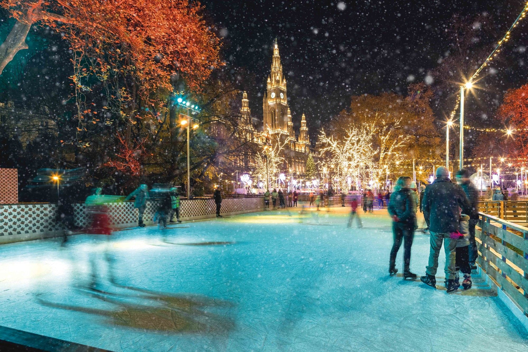 Ice skaters at the Vienna Ice Dream in front of the festively lit City Hall, surrounded by sparkling trees and falling snow.