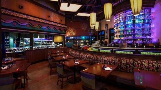 A restaurant dining area with tables, chairs, booths, and a bar in the background.