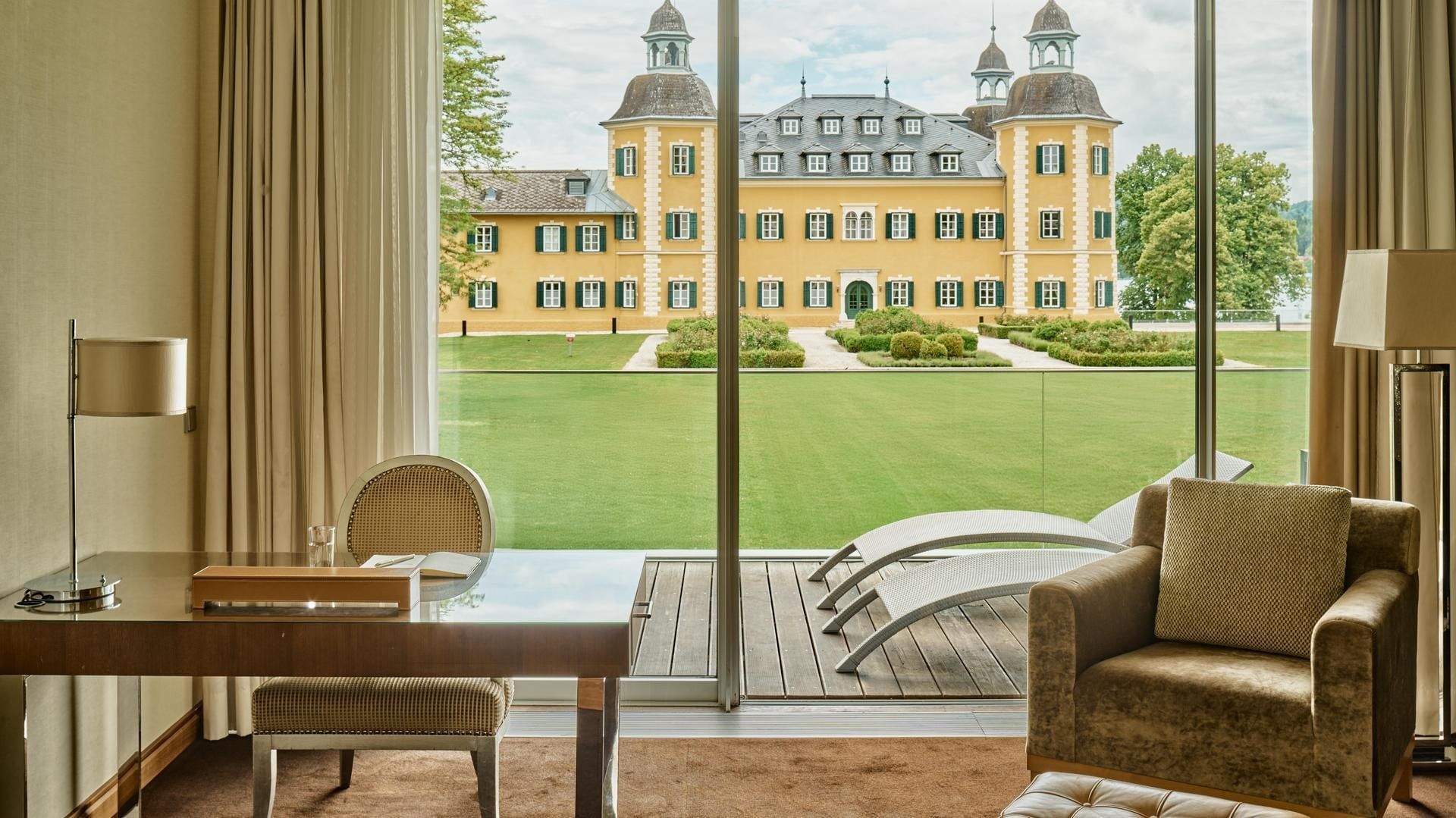 Room with desk, chair, and lounge chair by glass doors showing a large lawn and yellow building.