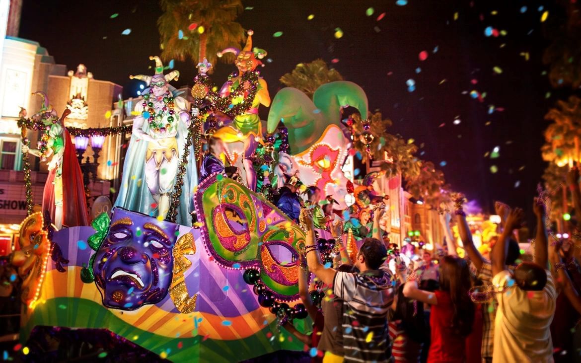 A large parade float with drama masks, a masquerade mask, jester masks, and jester figures glides through the street during a nighttime parade surrounded by excited people and confetti.