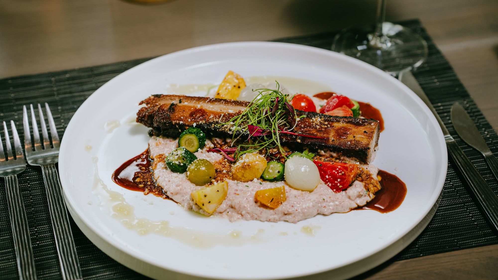 Plate with a meat dish, mashed potatoes, assorted vegetables, and sauce, accompanied by cutlery and a wine glass.