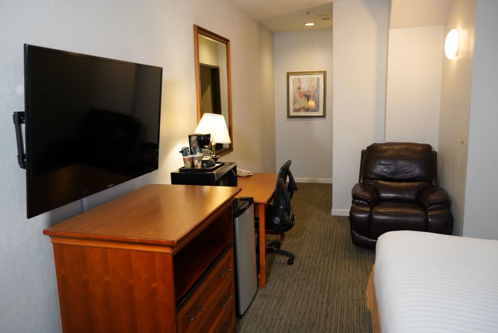 A wide view of a hotel room with a desk, TV, chair, recliner, and bed.