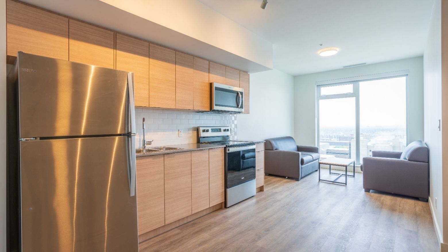 A view of a room with a sleek, full sized kitchen and sitting area with two couches close to large windows.