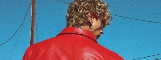Man with curly blonde hair in a red leather jacket stands against a blue sky.