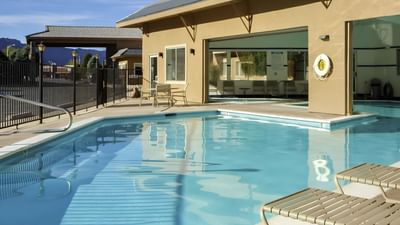 Sunny pool area at Moab Valley Inn with clear water, lounge chairs, a safety lifebuoy, and fenced surroundings