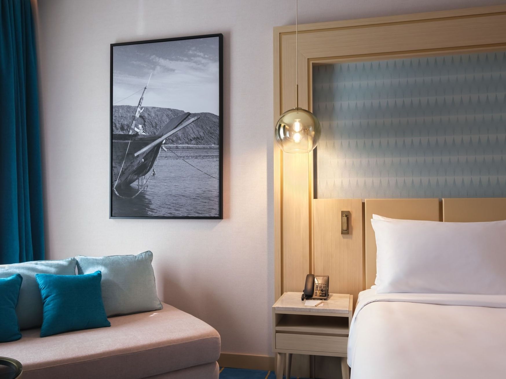 Modern hotel room with bed, couch, wall art of a boat, and a blue curtain.