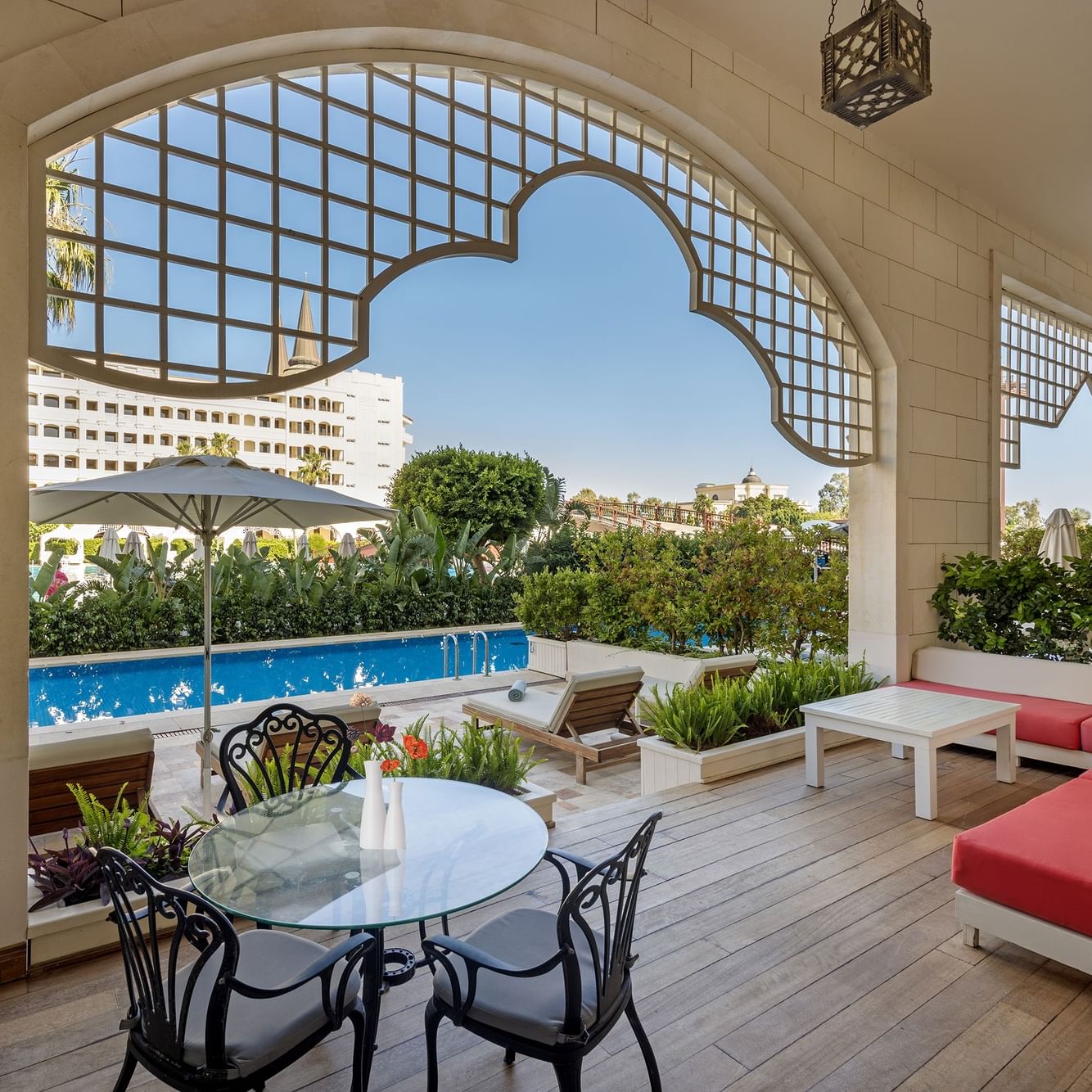 Veranda der First-Class Pool Duplex Suite im Titanic Mardan Palace mit Blick auf den Pool, umgeben von Grün, ausgestattet mit Tisch, Stühlen, rotem Sofa und Sonnenschirmen.