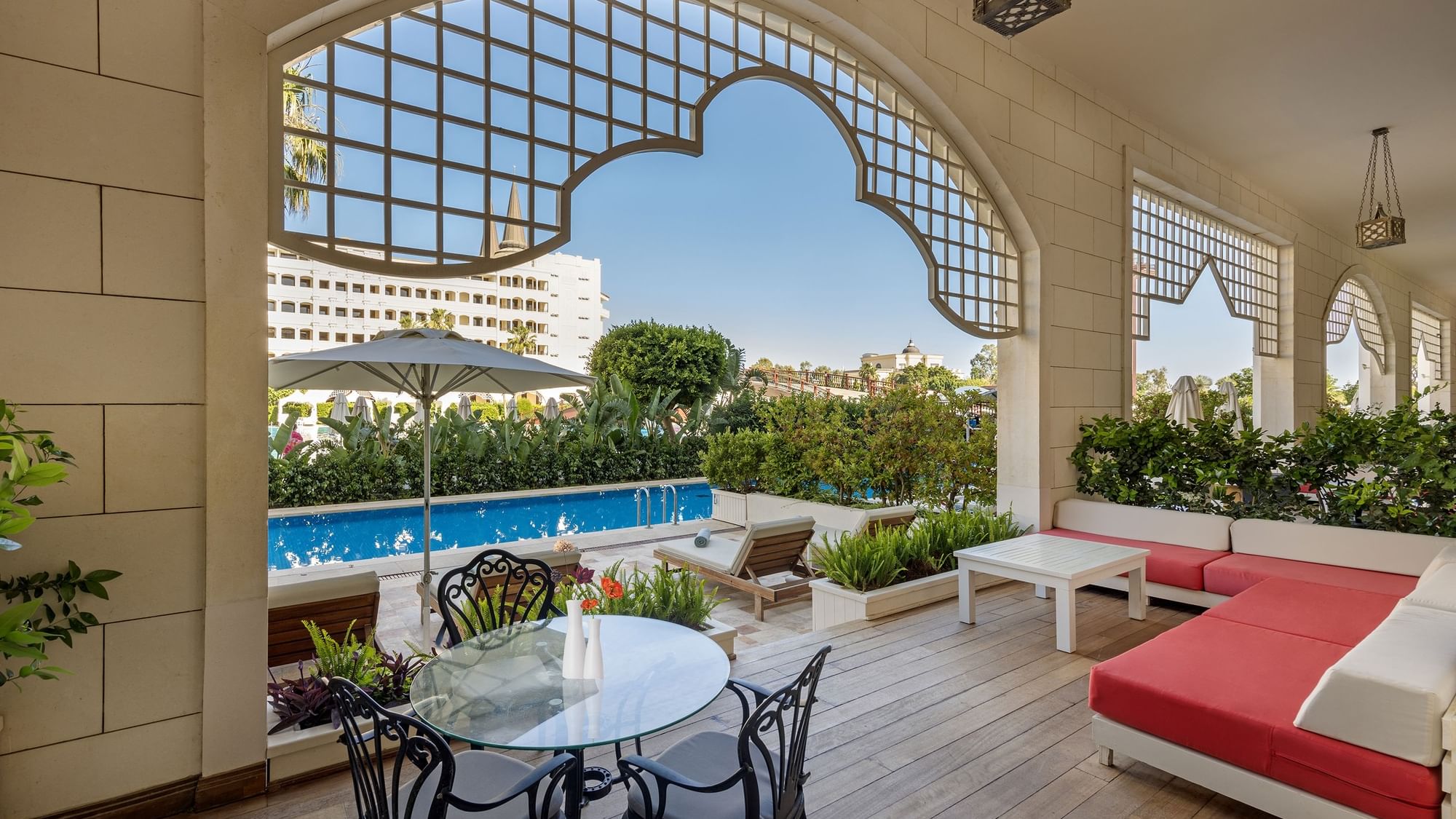 Veranda der First-Class Pool Duplex Suite im Titanic Mardan Palace mit Blick auf den Pool, umgeben von Grün, ausgestattet mit Tisch, Stühlen, rotem Sofa und Sonnenschirmen.
