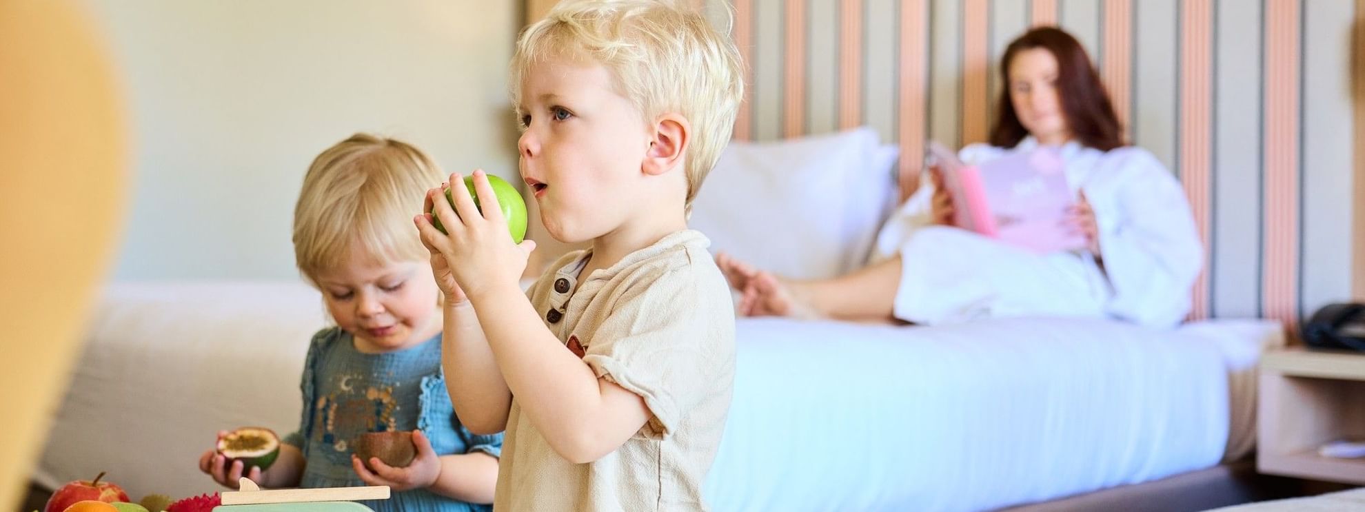 Two children eating fruit in a hotel room with a woman on a bed in the background.