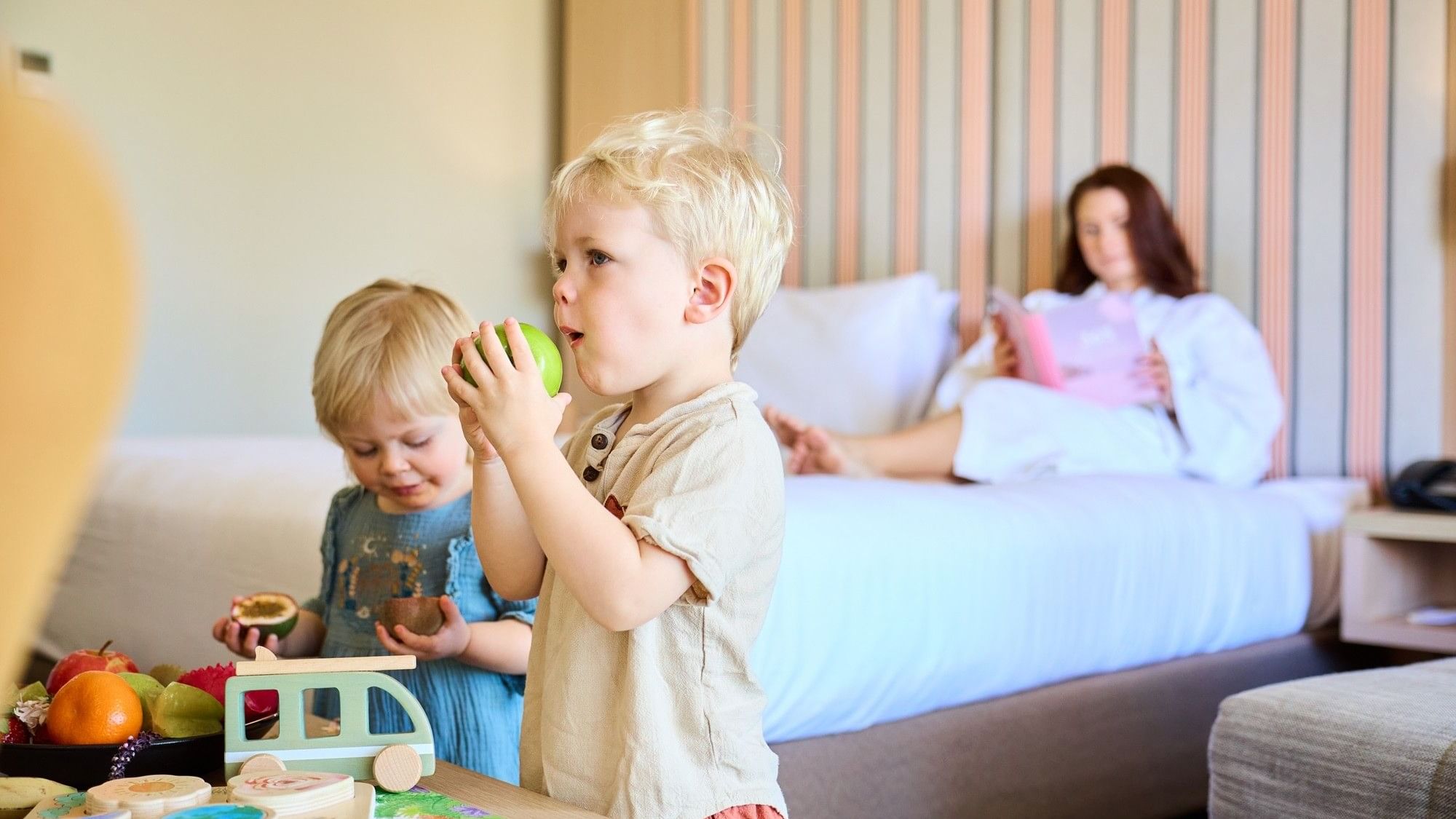 Two children eating fruit in a hotel room with a woman on a bed in the background.