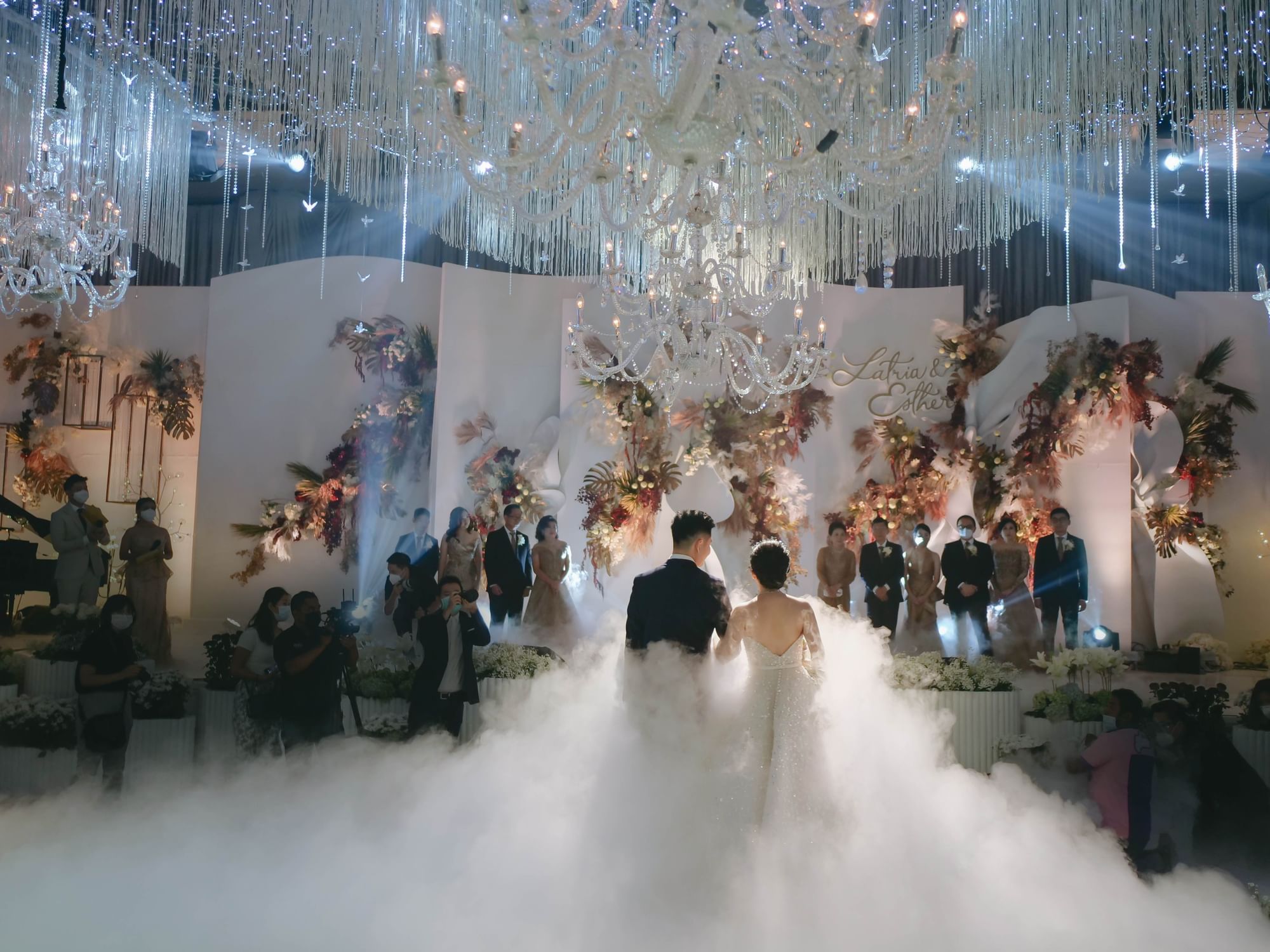 A couple stands together in a fog-filled ballroom, surrounded by elegant chandeliers, and floral decorations at Vasa Hotel Surabaya