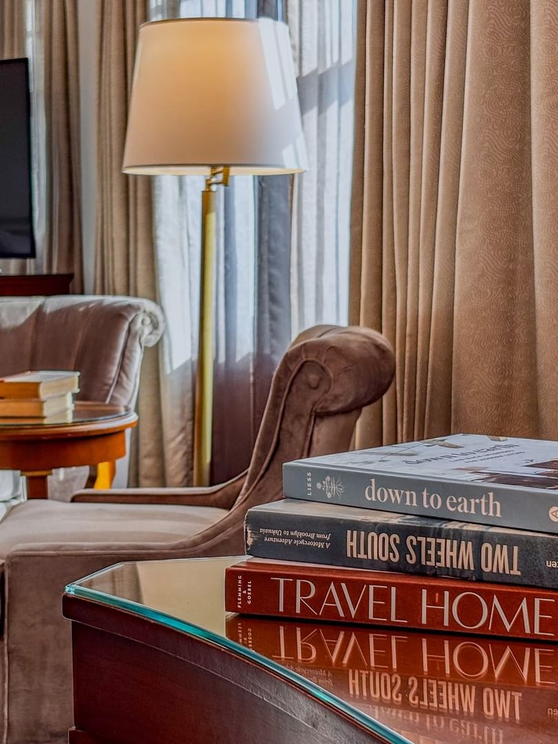 Stack of travel books on a glass table by velvet armchairs under a floor lamp in Deluxe King at Warwick Melrose Dallas