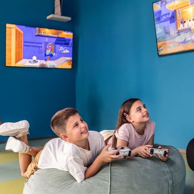 Due bambini giocano con console su un pouf in una stanza con pareti blu e due monitor.