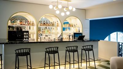 A bar with marble counter and four black stools, arched shelving with liquor bottles, and blue wall.