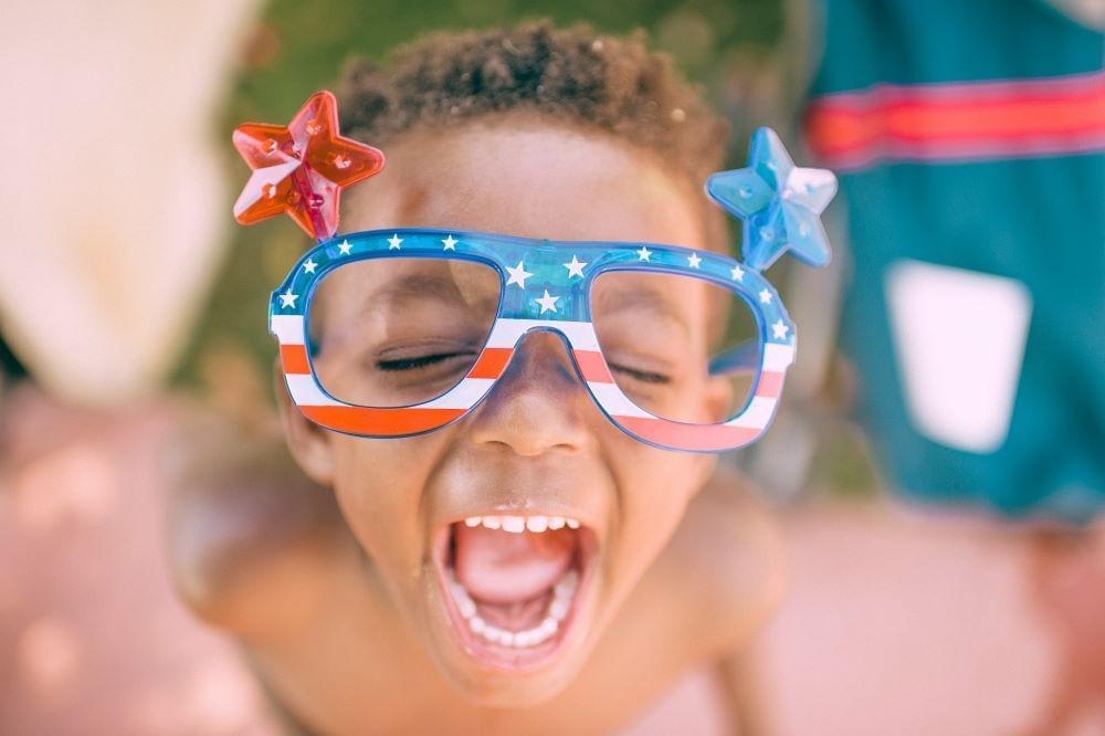 A young boy with curly hair wears patriotic red, white, and blue sunglasses with stars and stripes, his eyes closed, and mouth wide open.